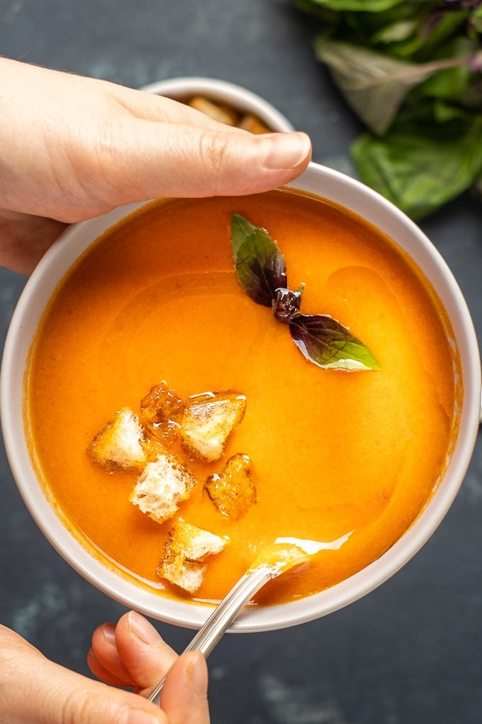 Woman holding a white bowl of tomato soup with fresh tomatoes topped with croutons and fresh basil leaves.