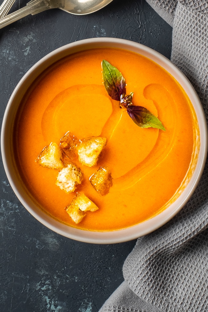 Homemade tomato soup topped with croutons and fresh basil leaves in a white bowl on a dark background.