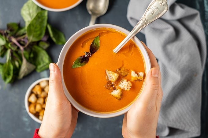 Hands holding a bowl of tomato soup with fresh tomatoes.