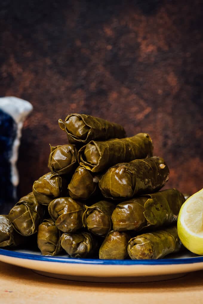 A stack of stuffed grape leaves on a plate, half lemon and yogurt sauce on the side.