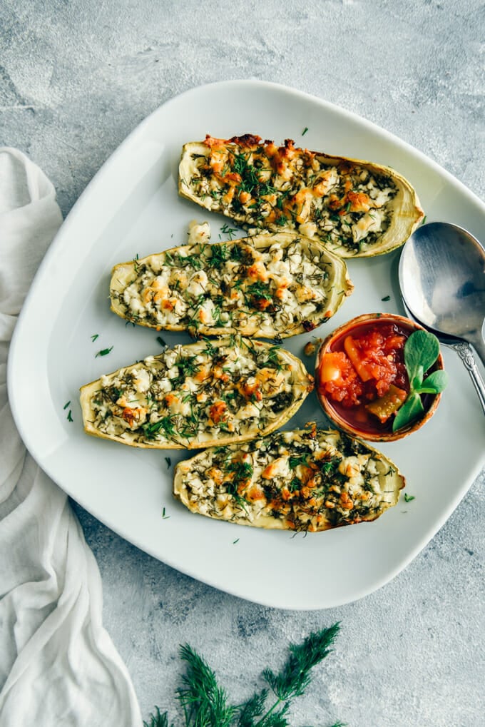 Zucchini boats stuffed with a cheese filling served on a white plate with a chili sauce and a spoon on the side