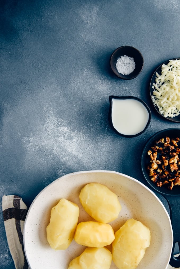 Cooked and peeled potatoes in a white ceramic bowl for cheesy mashed potatoes on a dark background.