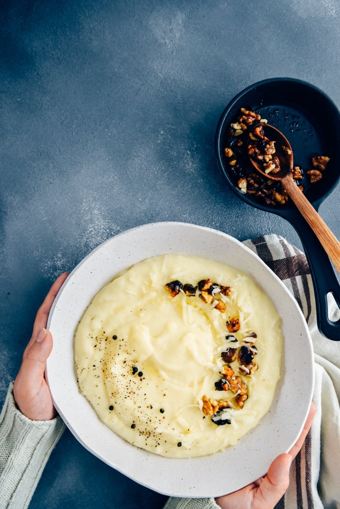 Hands holding cheesy mashed potatoes with roasted walnuts and black pepper in a white ceramic bowl