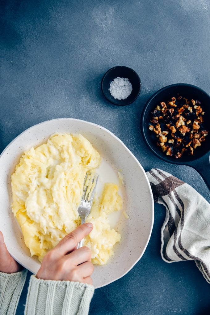 Hands mashing potatoes in a white ceramic bowl using a fork.