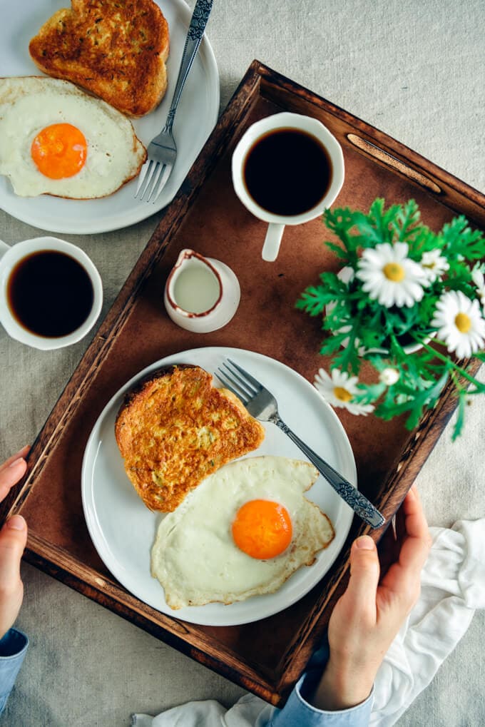 Woman holding a wooden tray with a French toast and a fried egg on a white plate and a cup of coffee, milk and flowers on the side. Another plate with french toast and an egg and another cup of coffee accompany out of the tray. This Easy French Toast recipe with a spicy twist makes your mornings worth waking up to. It is crispy on the outside and soft on the inside. Perfect with both savory and sweet toppings but I love it as it is.