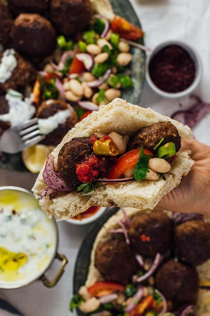 A hand holding a flatbread sandwich with homemade meatballs, bean salad, sumac onions and pickles.