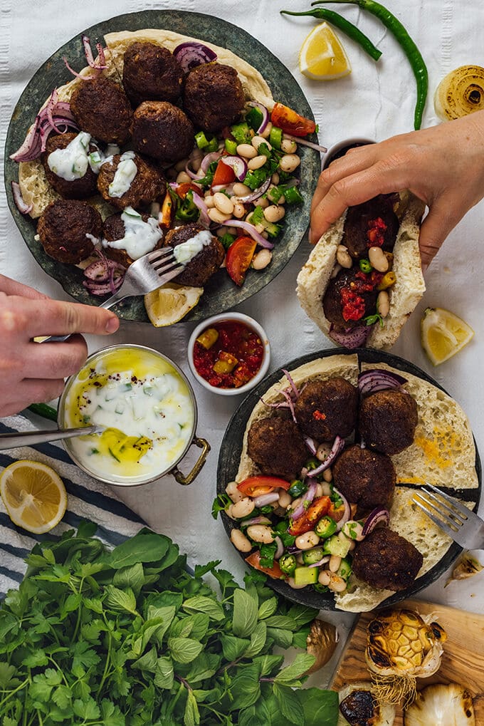 Hands grabbing authentic Turkish meatballs with a fork and in a sandwich, bean salad, sumac anions, Turkish cacik and lemon wedges accompany.