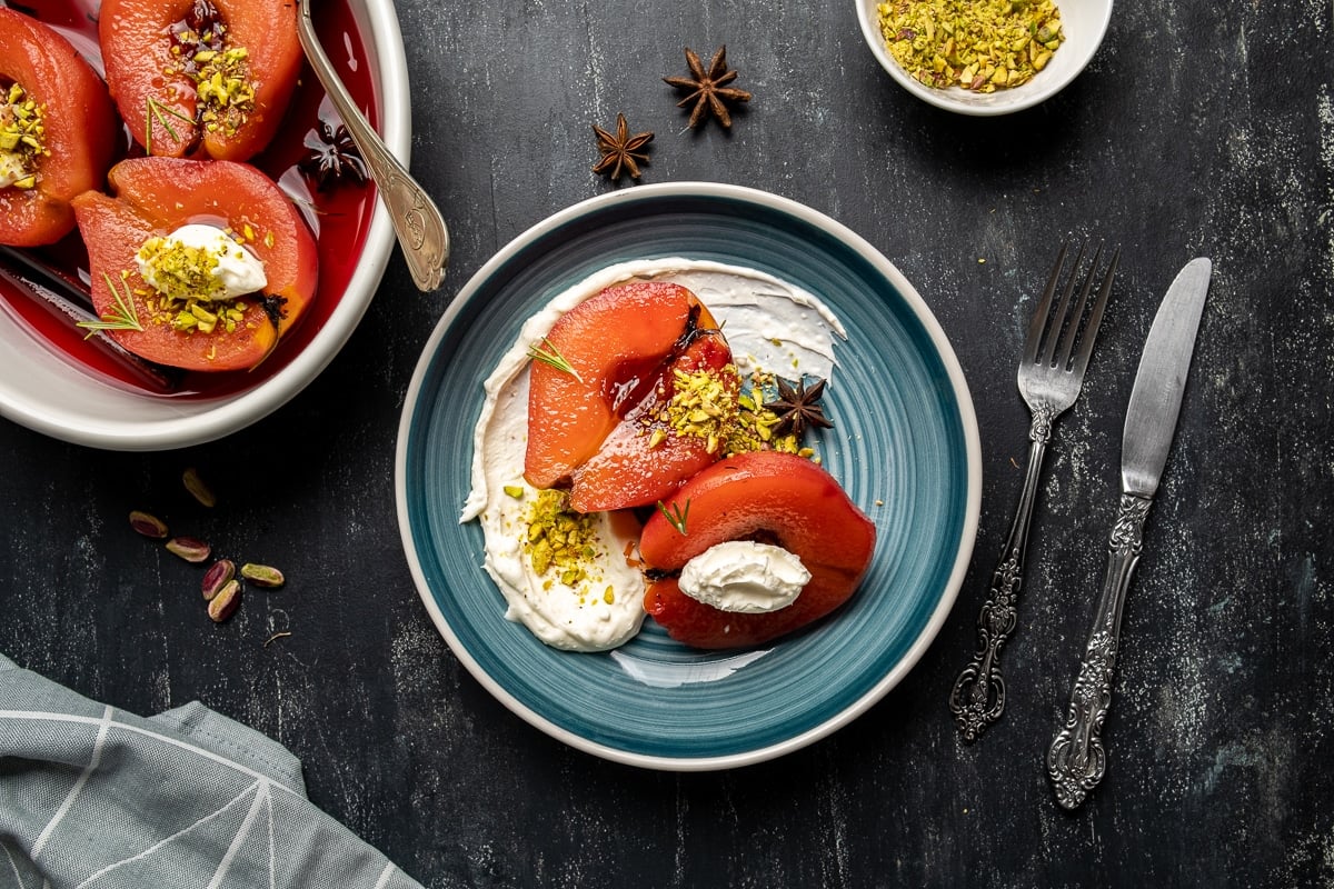 Poached quince fruit served on a plate with pistachios and clotted cream, a white pan with the rest of the quinces, a knife and a fork on the side.