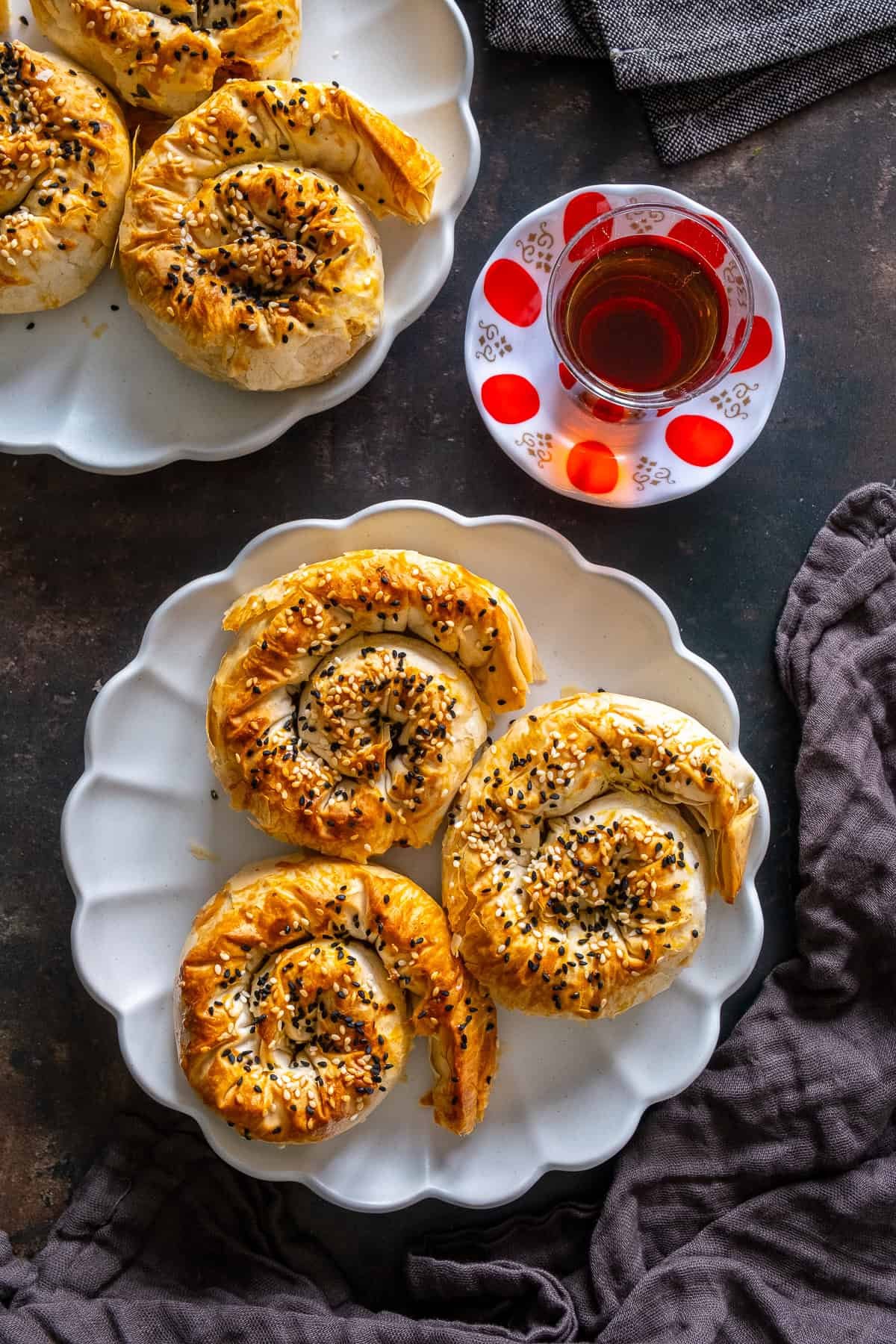 Top view of spiral potato böreks topped with sesame and nigella seeds on a white plate, served with Turkish tea.