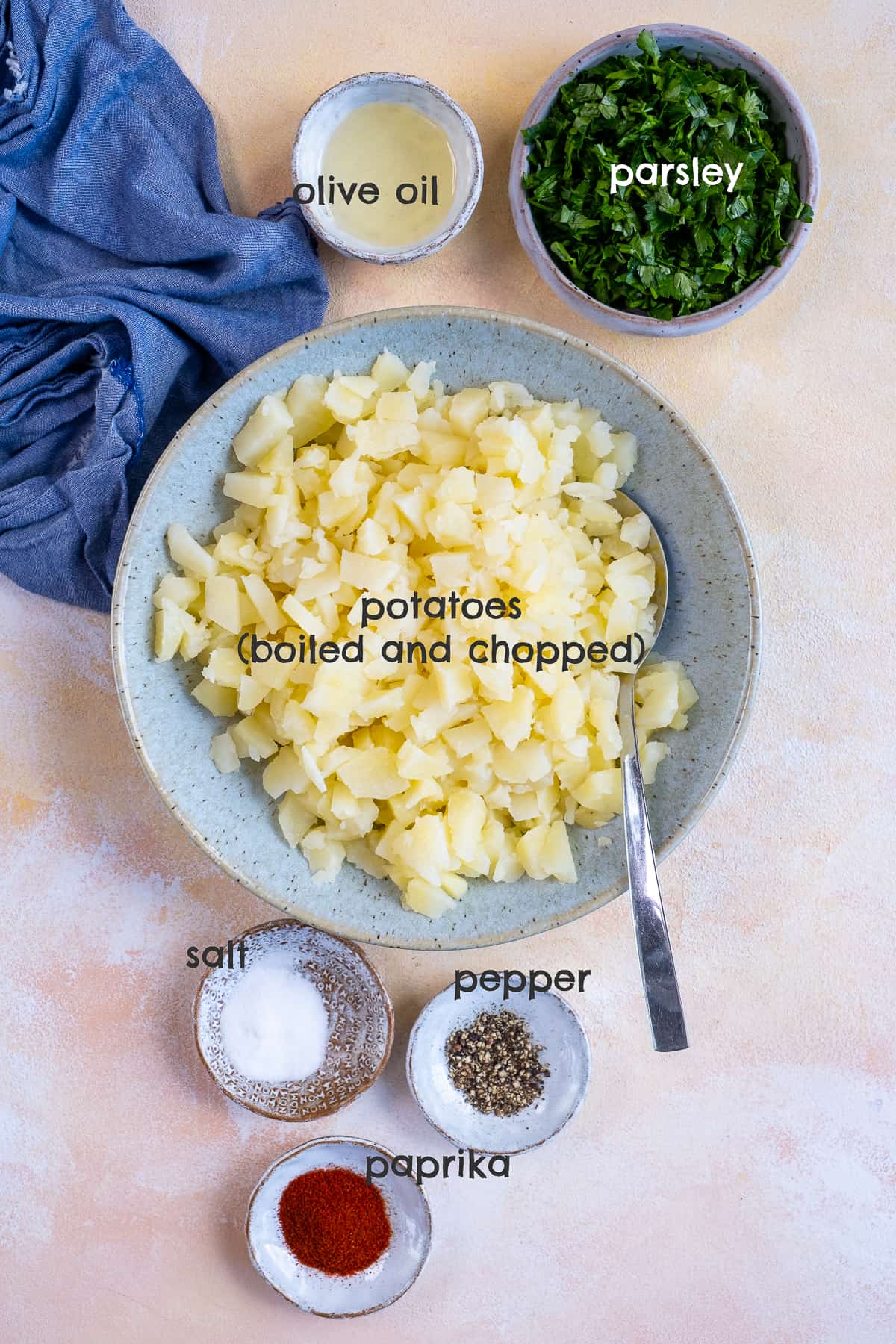 Chopped boiled potatoes in a bowl, parsley, olive oil, salt, pepper and paprika in separate bowls on a light background.