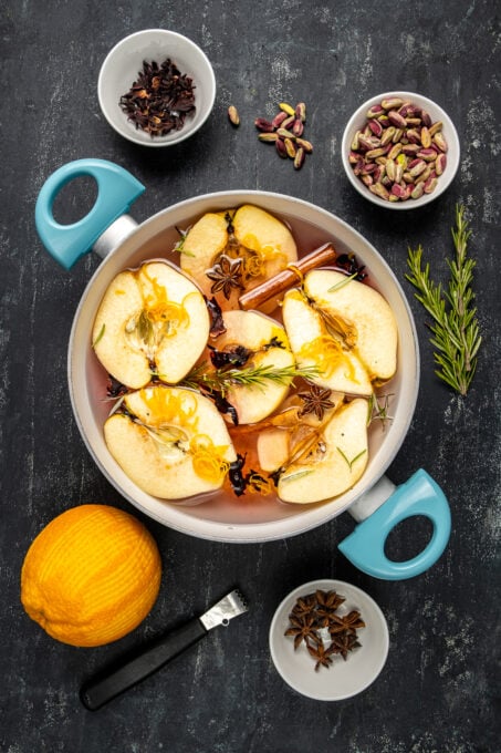 Peeled and halved quinces in a pot wih cinnamon sticks, rosemary leaves and star anise on a dark background.