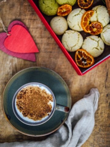 A cup of Turkish salep garnished with cinnamon powder photographed on a wooden background. Accompanied by red heart shaped ornaments, a box full of cookies, cinnamon sticks and orange chips.
