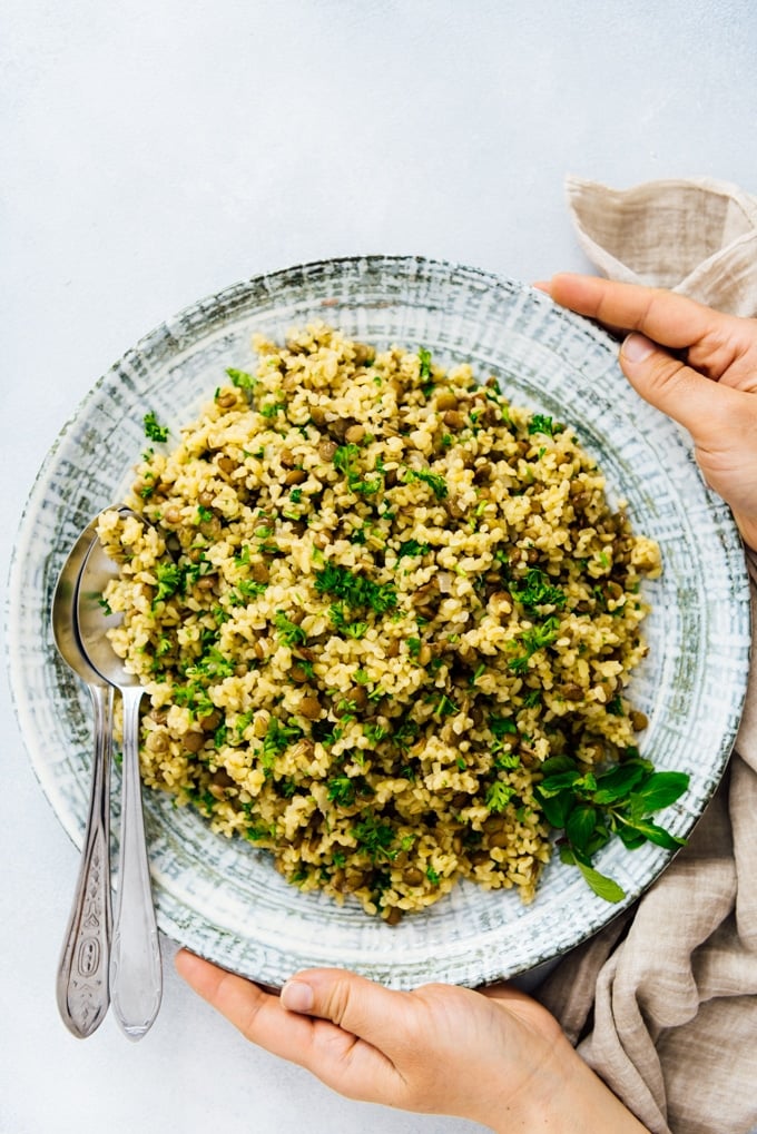 Woman serving bulgur wheat pilaf with green lentils and herbs in a large ceramic bowl.