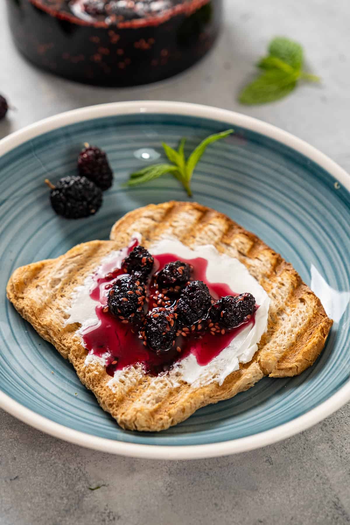 A slice of toasted bread topped with cream cheese and mulberry jam on a blueish plate, garnished with fresh mint and fresh mulberries.