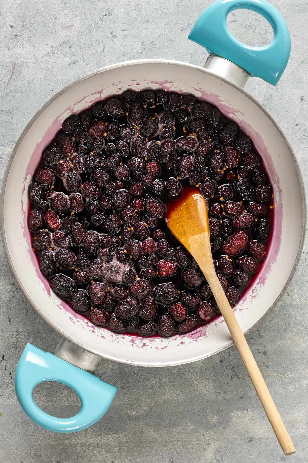 Mulberries cooking in a white pan and a wooden spoon inside it.