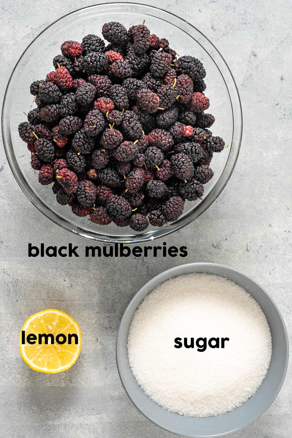 Fresh black mulberries in a bowl, half lemon and a bowl of sugar on a grey background.