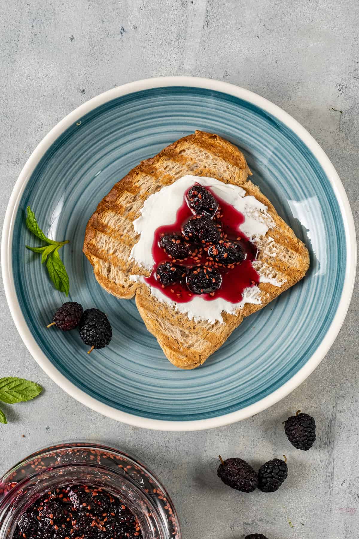 Toasted bread topped with cream cheese and mulberry jam on a blueish plate.