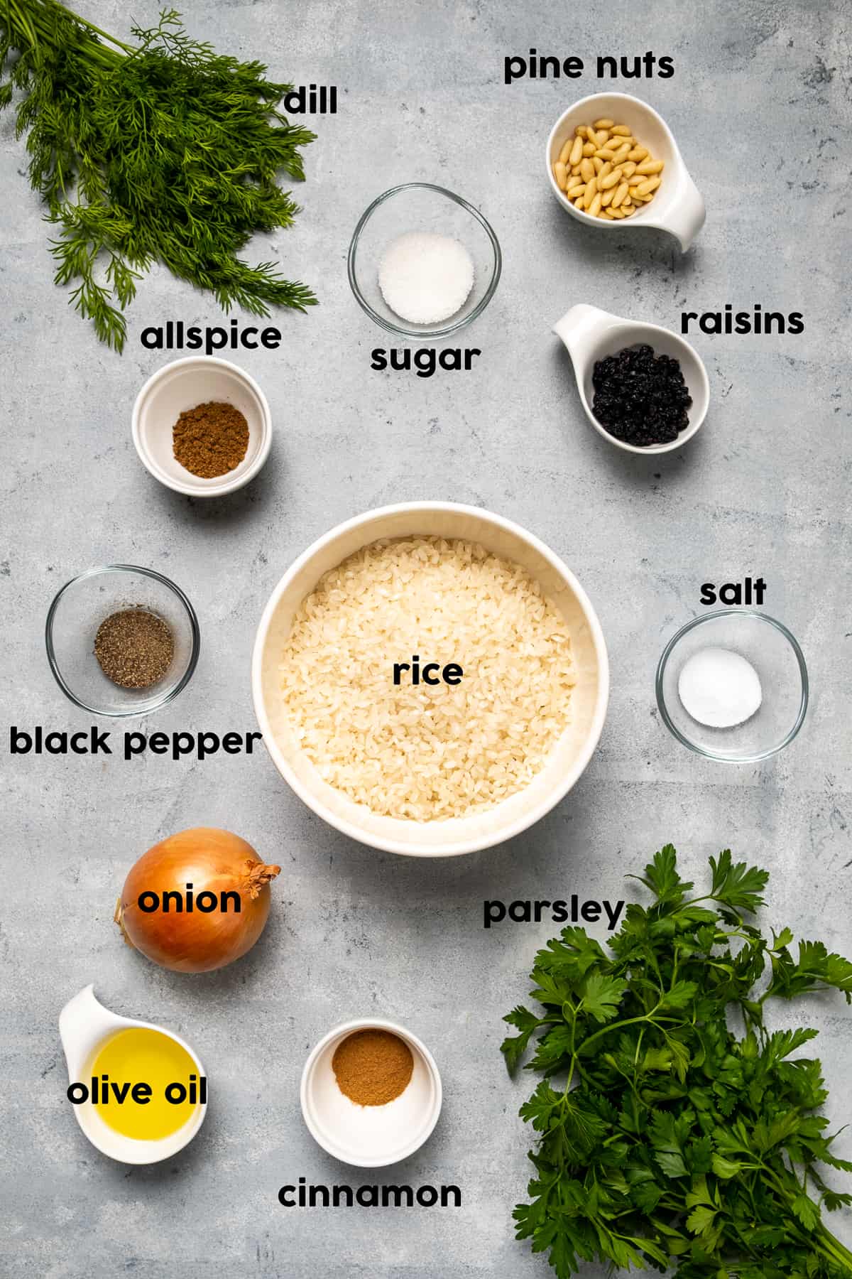 Rice in a large bowl, pine nuts, raisins, dill and parsley, spices, onion and olive oil on a grey background.