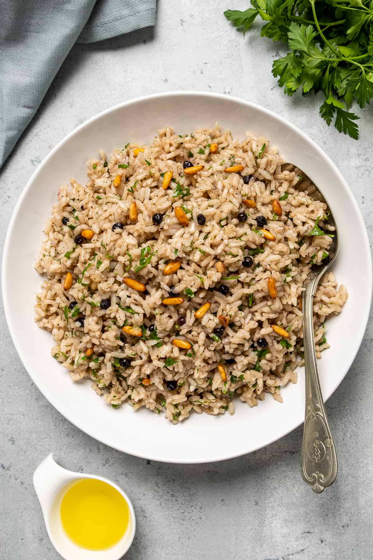 Spiced rice pilaf made with raisins, pine nuts and herbs served in a white bowl with a spoon inside it. Parsley and olive oil on the side.