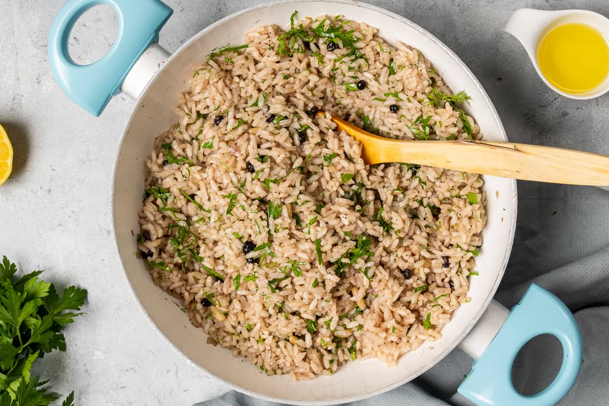 Rice with raisins and pine nuts garnished with parsley in a white pan and a wooden spoon in it.
