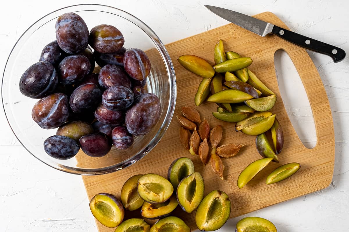 Chopped damson plums on a wooden board with a knife on the side and whole plums in a large glass bowl.