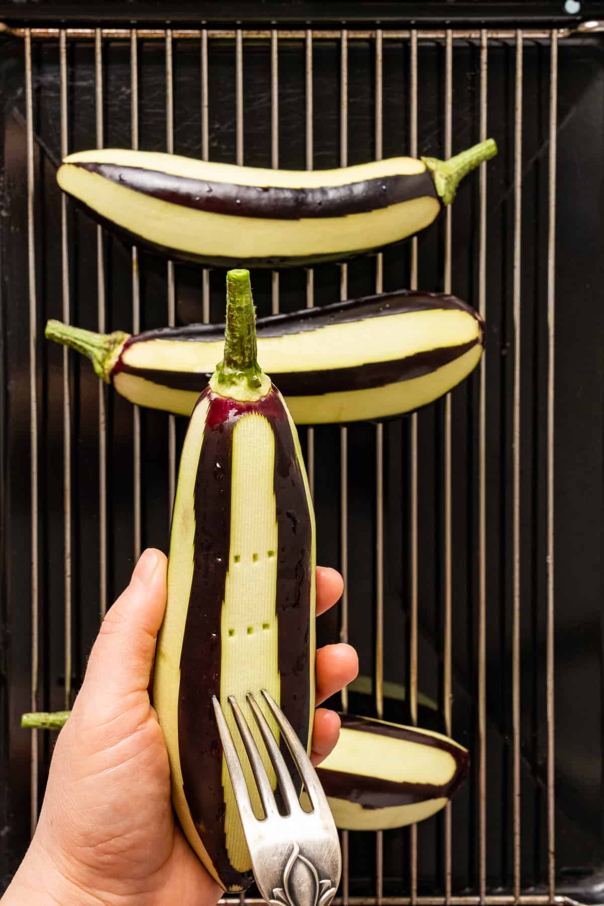 Hands pricking an eggplant with a fork and more eggplants on a grilling rack.