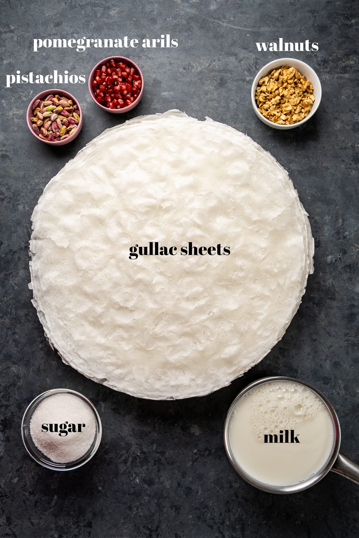 Gullac sheets surrounded by pistachios, pomegranate arils, walnuts, sugar and milk in separate bowls on a dark background.