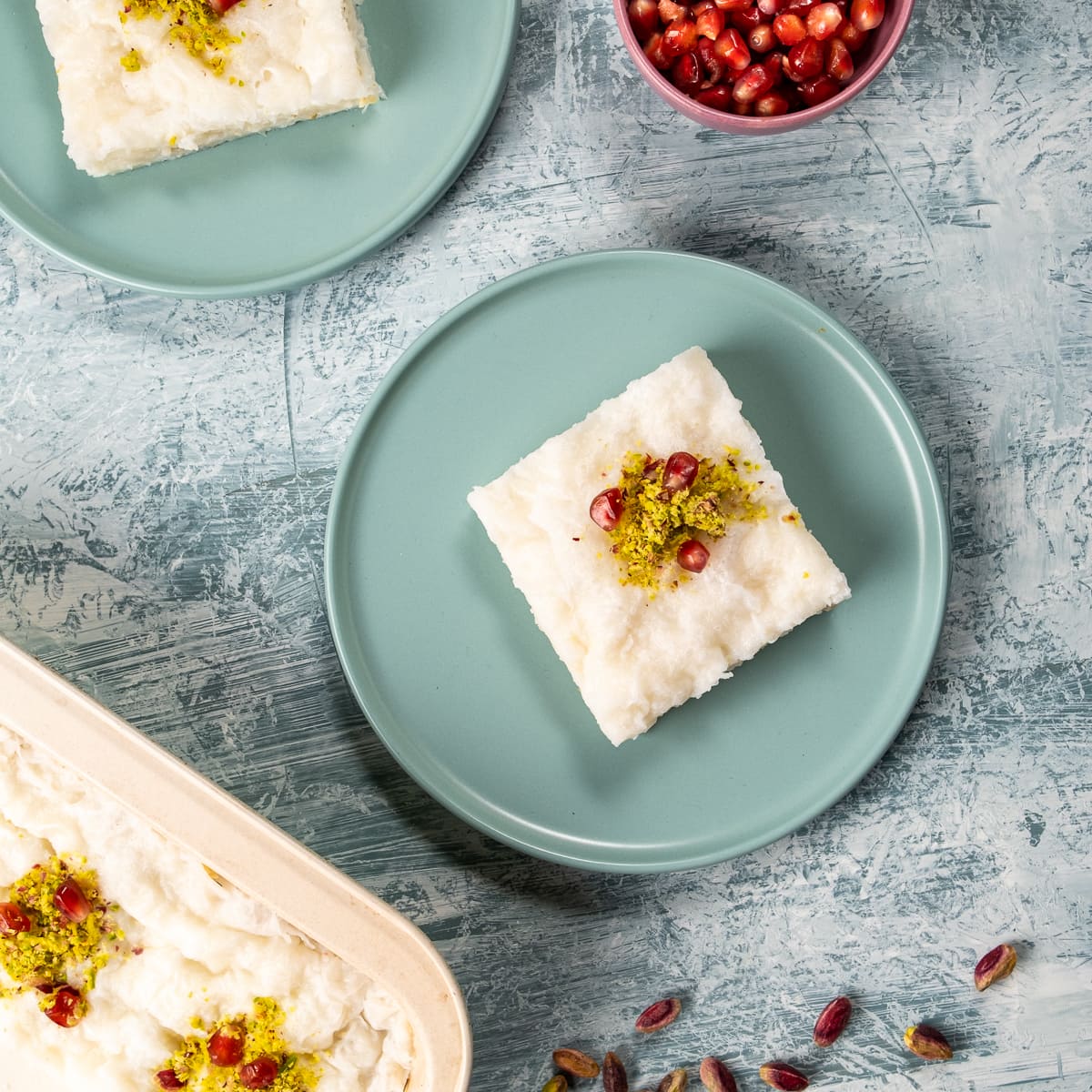 A slice of gullac dessert on two pastel colored plates on a light background, a bowl of pomegranate arils on the side.