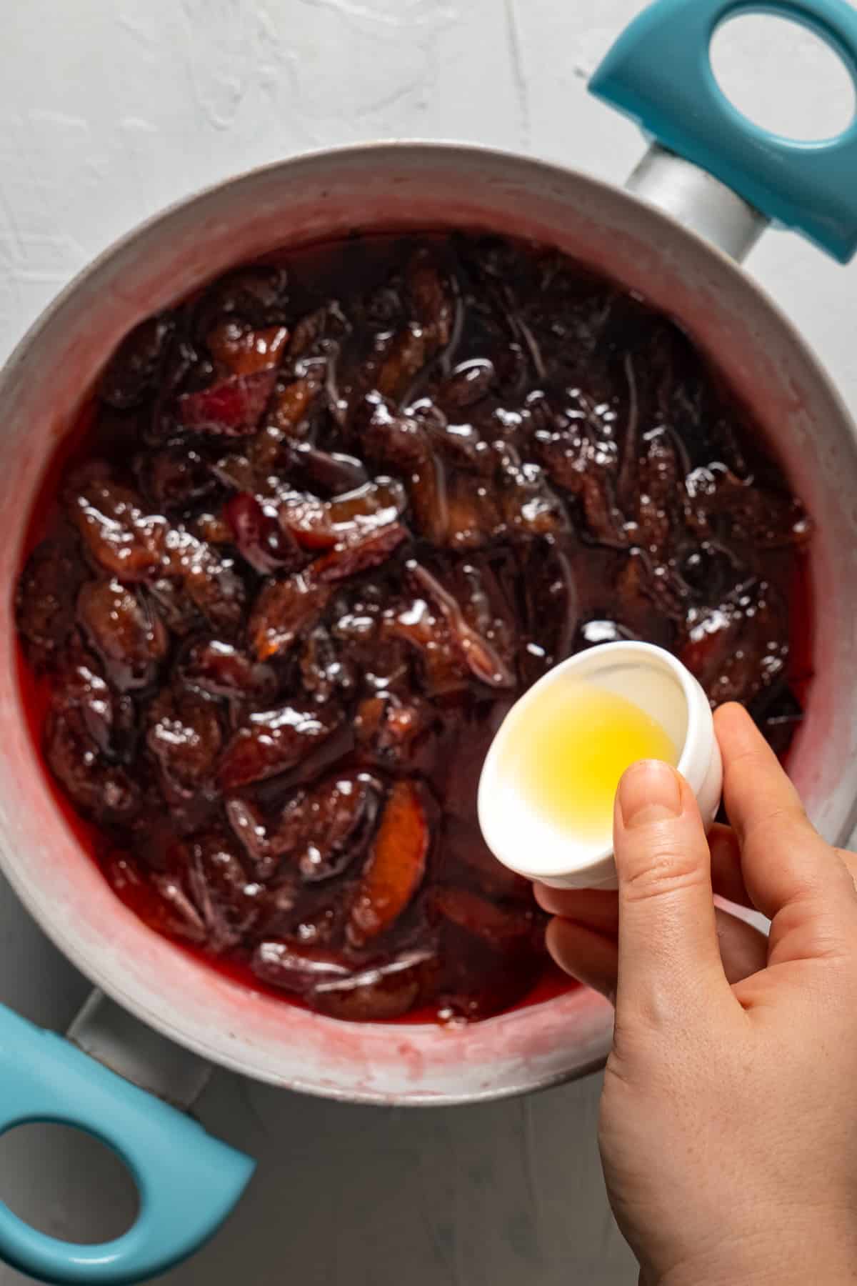 A hand pouring lemon juice with a small white bowl into the cooking plum jam.