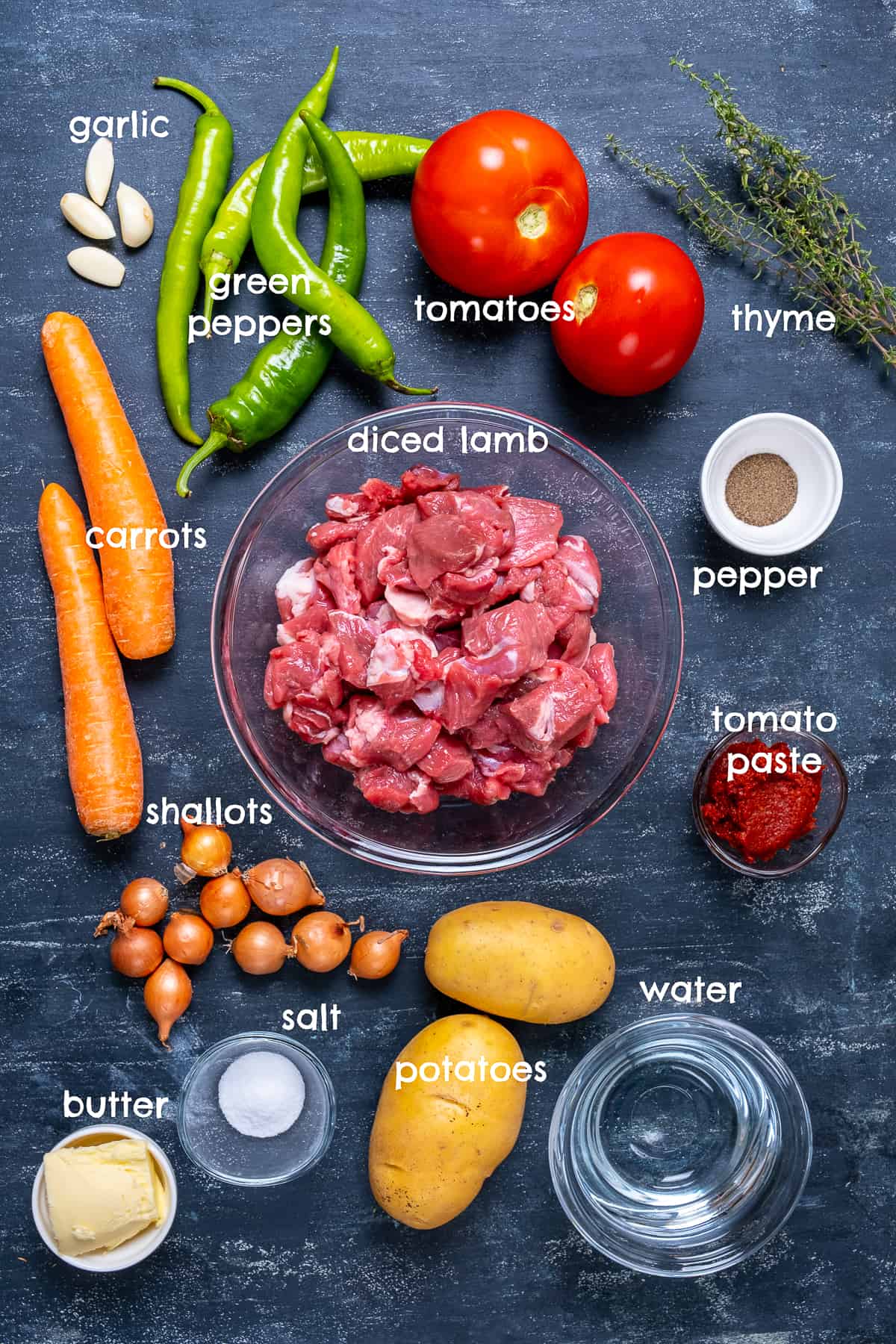 Diced lamb, tomatoes, peppers, carrots, shallots, potatoes, garlic cloves, fresh thyme, butter, tomato paste on a dark background.