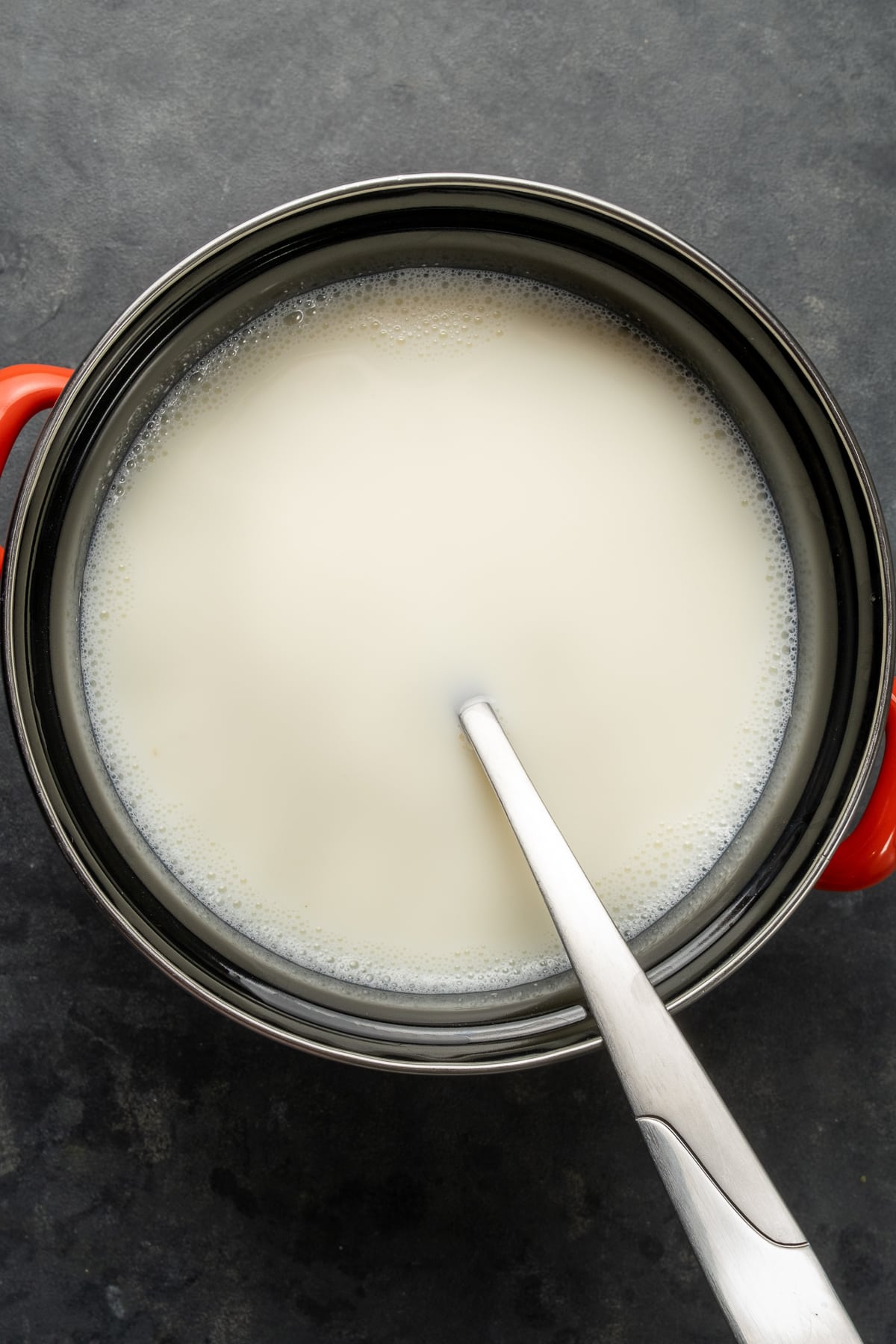 Warm milk in a pot and a spoon inside it on a dark background.