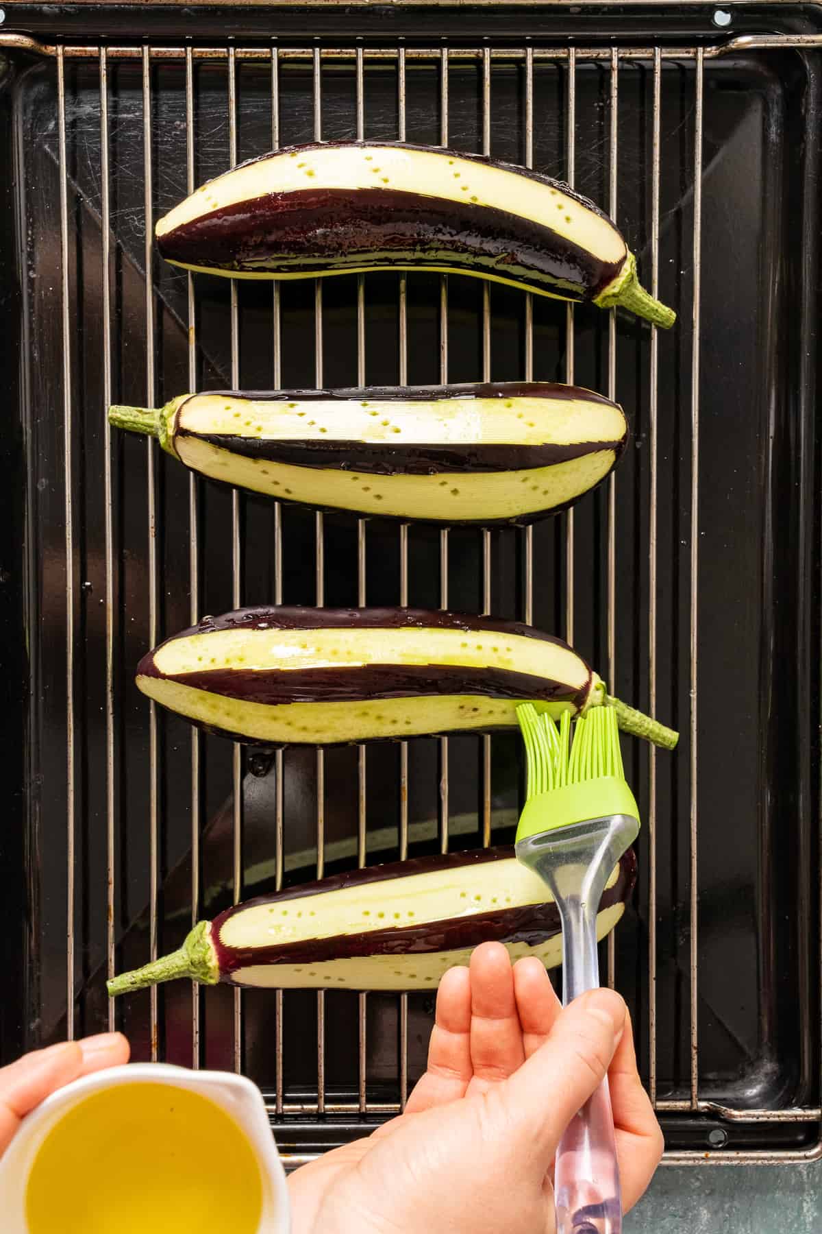 Hands oiling eggplants on a grilling rack with a brush.