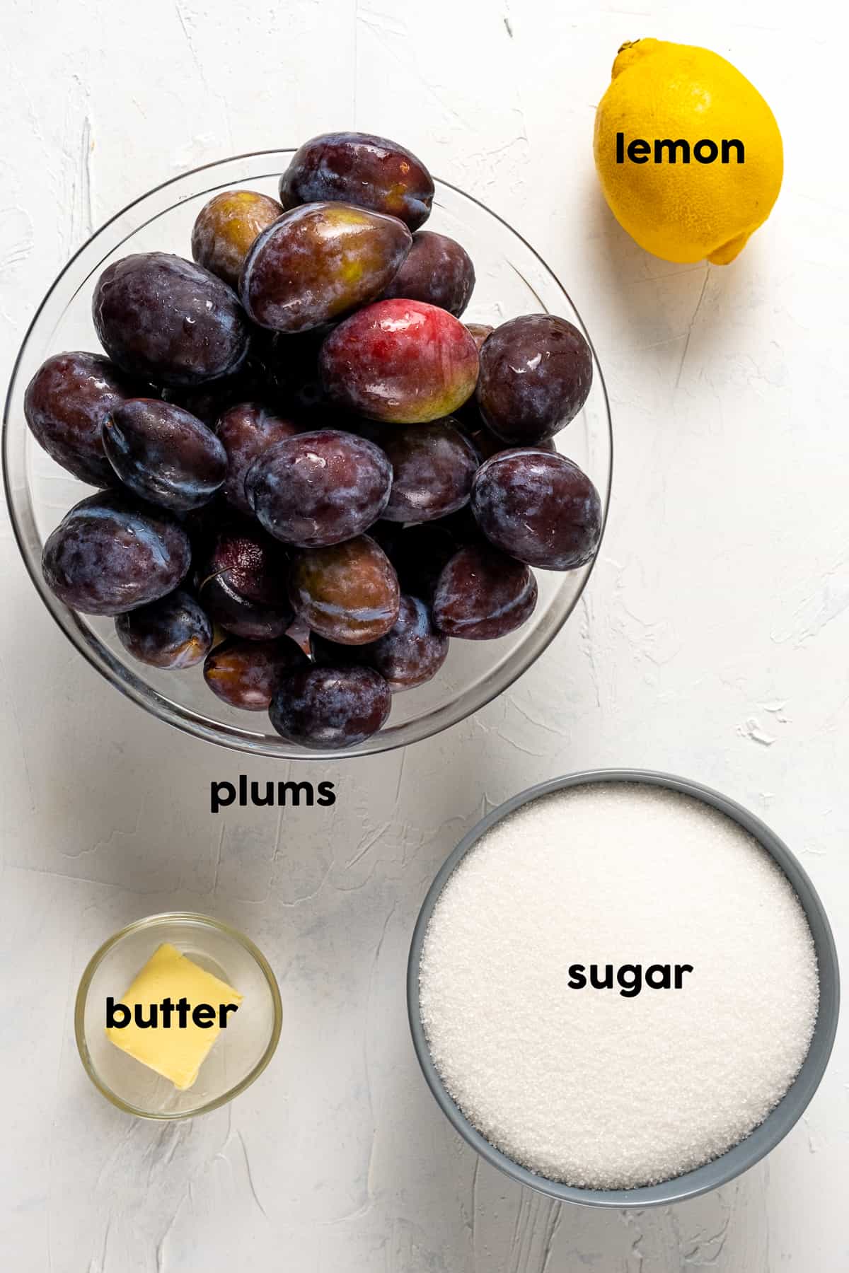 Fresh plums in a large bowl, lemon, sugar and butter on the side.