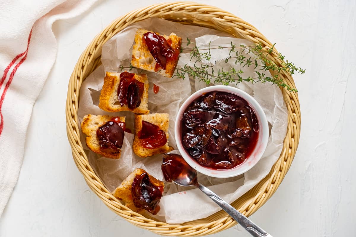 Plum jam served in a small white bowl with toasted bread bites topped with jam all in a basket.