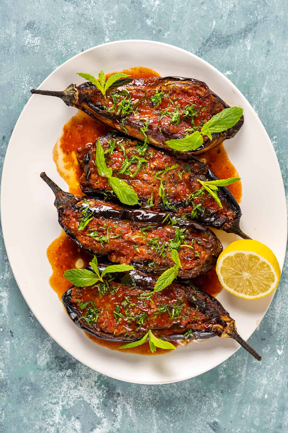 Imam bayildi eggplants served with tomato sauce, herbs and a half lemon in a white oval dish.