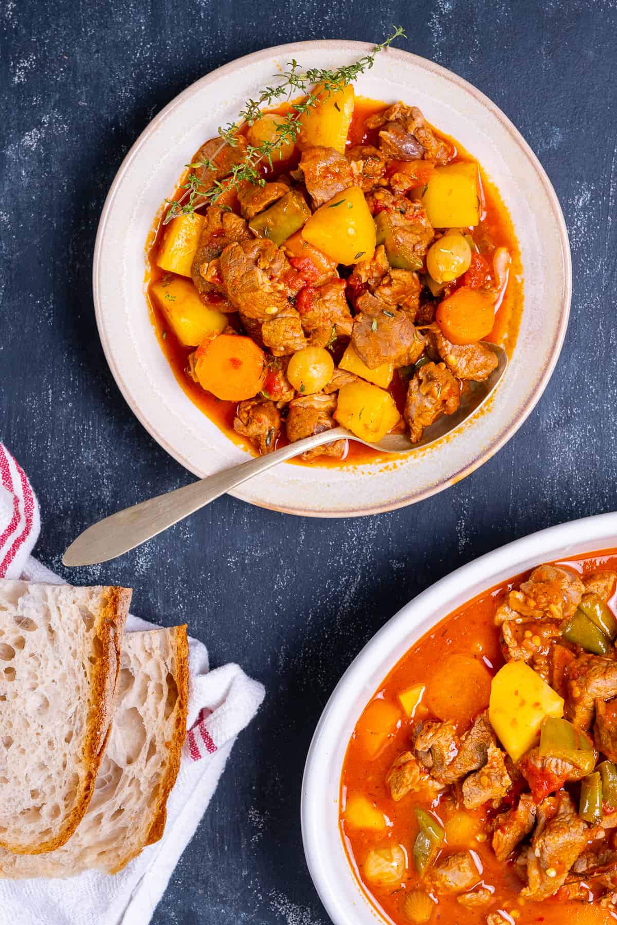 Lamb stew with potatoes and carrots in a large bowl and a spoon in it. Bread slices and another dish with stew on the side.