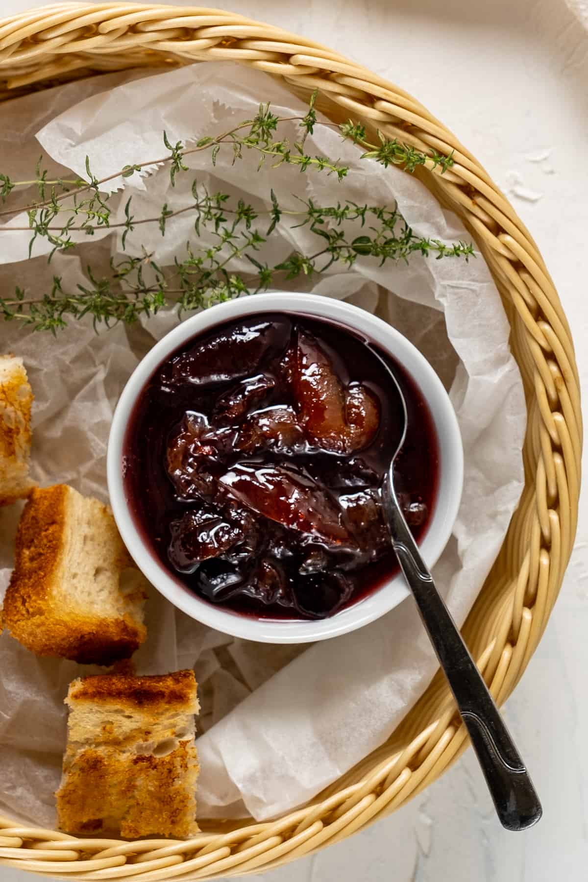 Plum jam in a small white bowl with a spoon in it and toasted mini bread slices and fresh thyme all in a basket.