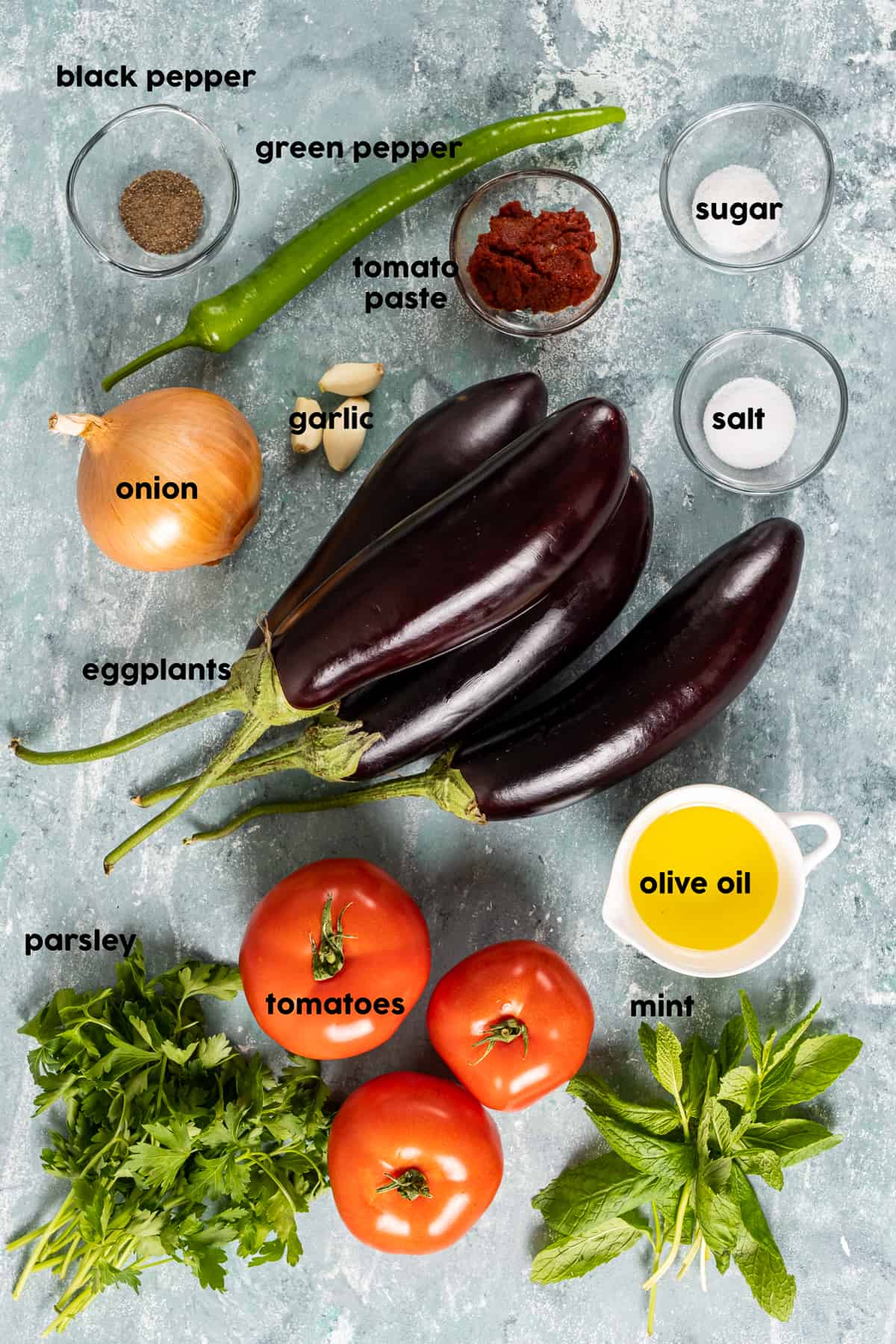 Eggplants, a green pepper, an onion, a bunch of parsley and mint, olive oil, garlic, tomato paste and spices in mini bowls on a grey background.
