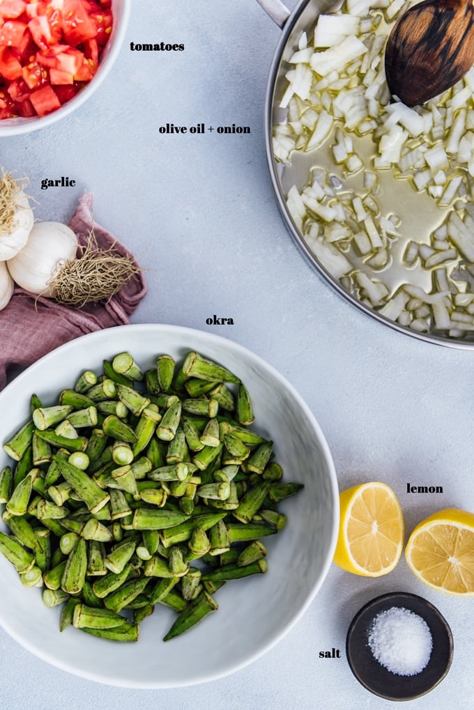 Chopped onions in a pan, lwhole garlic, halved lemon, fresh okra and tomatoes on a light background.