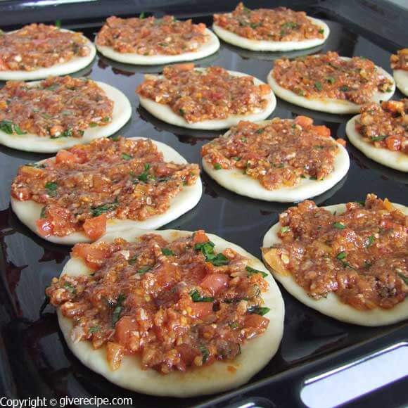 Mini lahmacun discs topped with a mixture of ground meat, tomatoes and spices on a baking sheet.