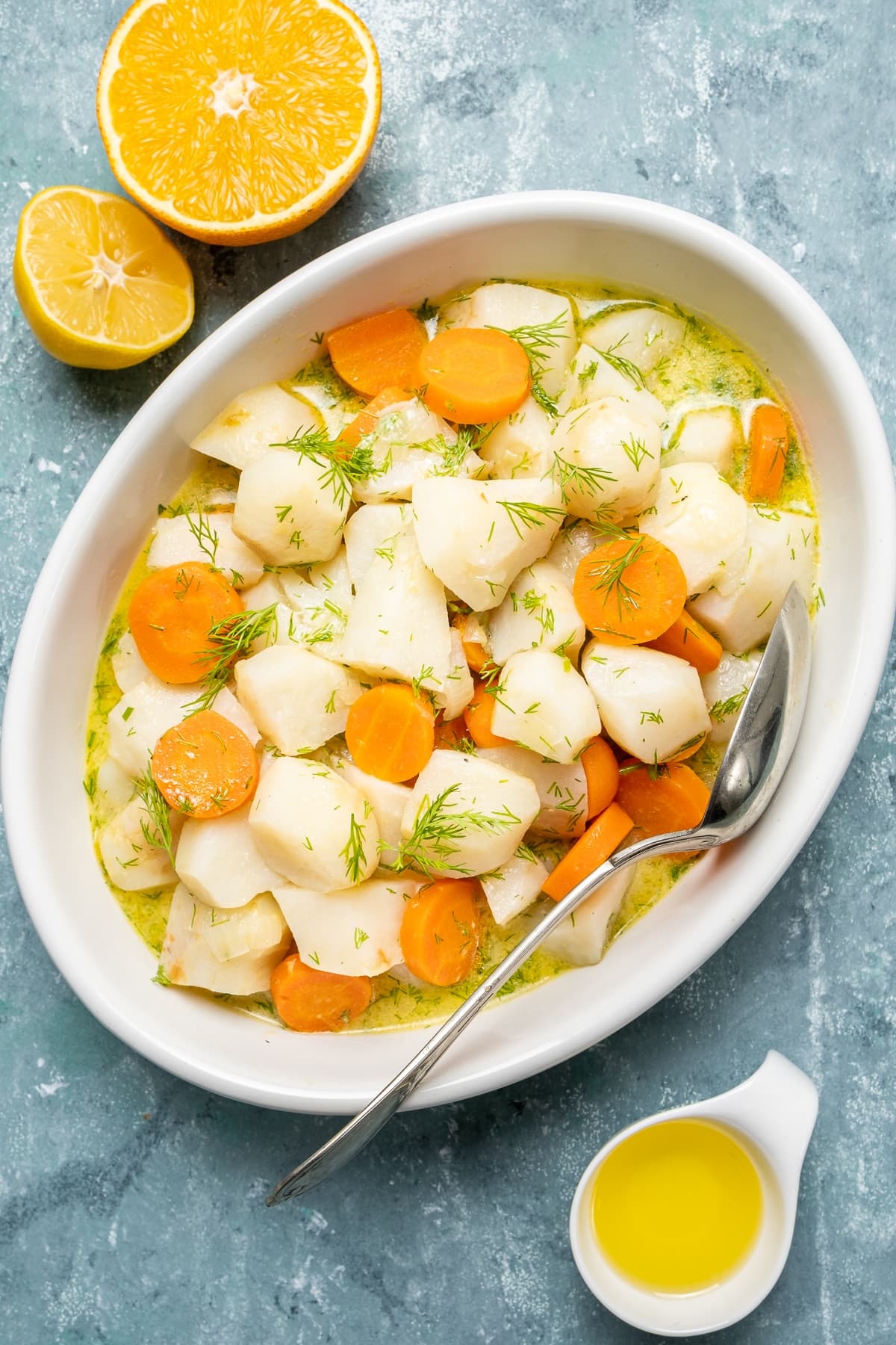 Braised sunchokes and carrots garnished with fresh dill in a white oval bowl and a spoon inside it. An orange cut into two on the side.