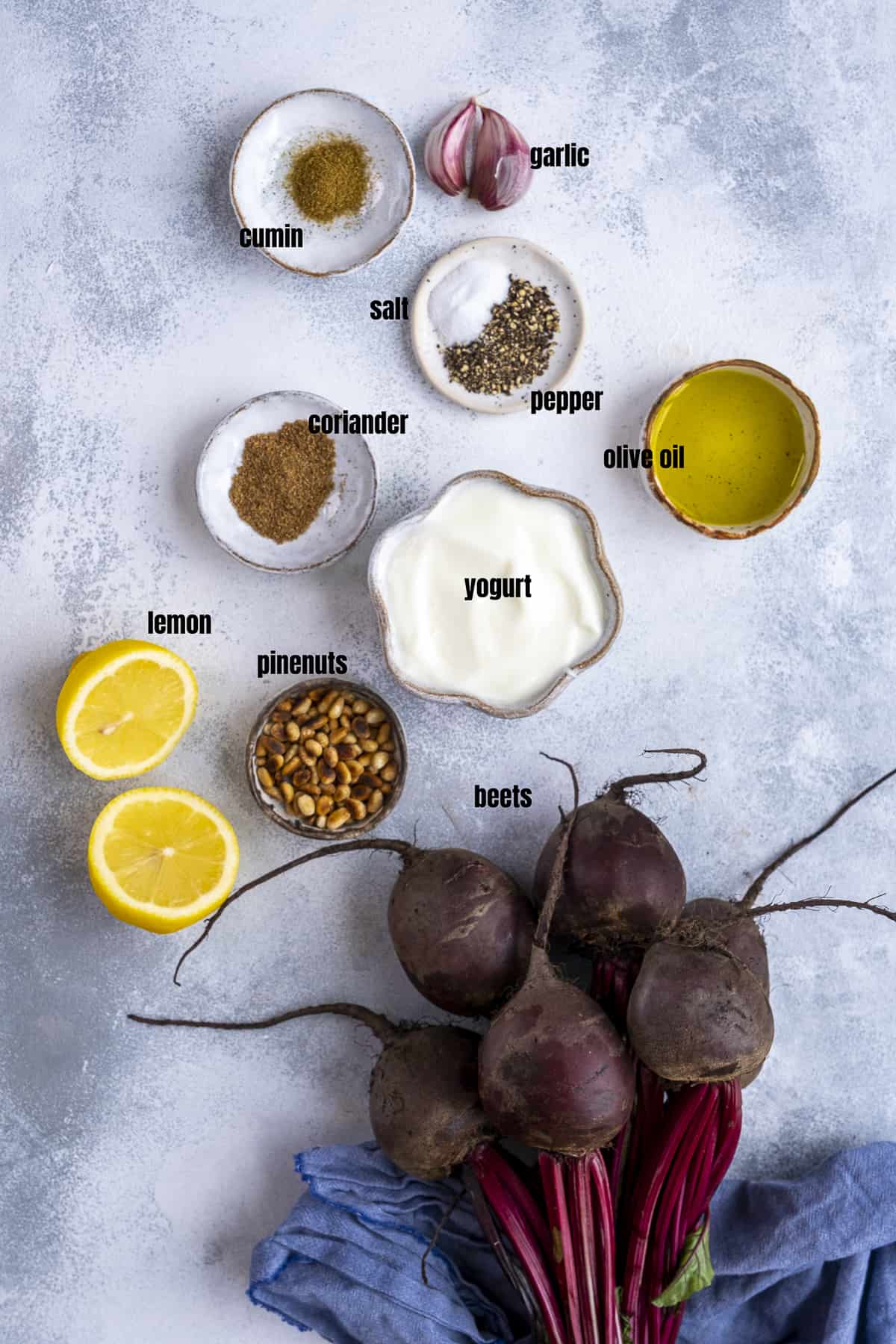 A bunch of beets, halved lemon, yogurt, garlic cloves, olive oil, cumin, coriander, salt and pepper, toasted pine nuts on a grey background.