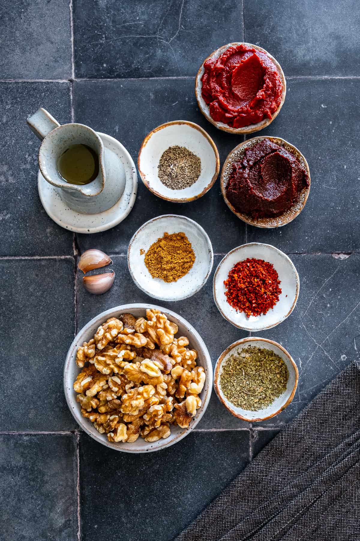 Ingredients for acuka arranged on a countertop, including walnuts, tomato paste, pepper paste, garlic, olive oil, cumin, pul biber, black pepper and oregano.