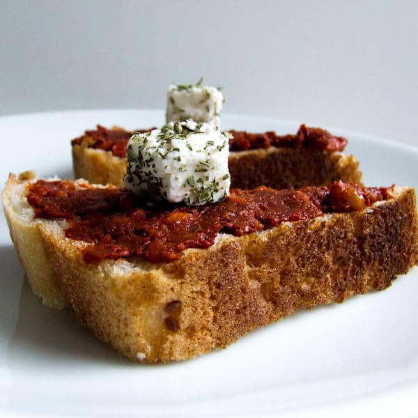 Acuka spread slathered on a piece of bread and topped with a cube of herbed cheese.