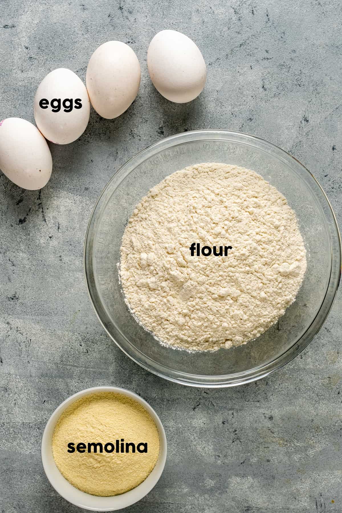 Flour in a large mixing bowl, semolina flour in a small bowl and eggs accompany on a grey background.