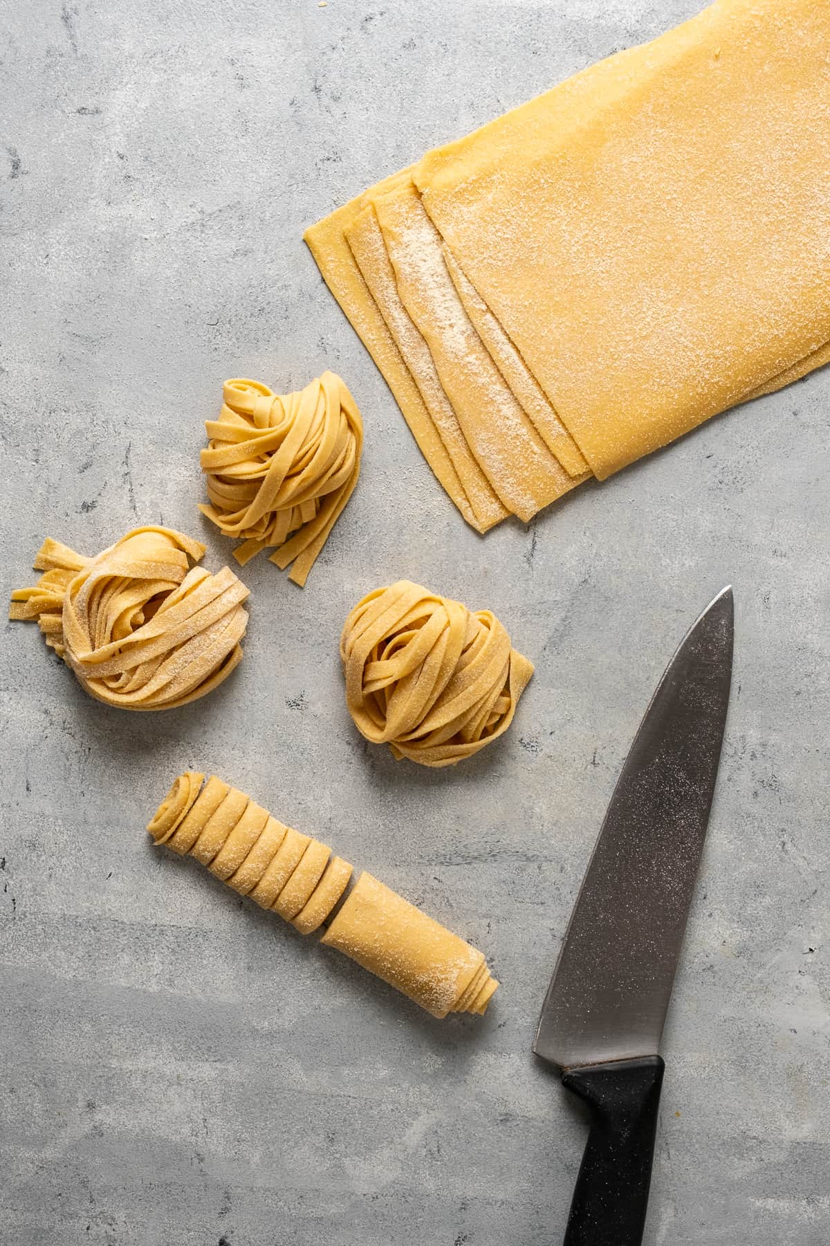 Pasta dough sheets piled, one sheet rolled and cut, three nests of dough strips and a knife on a grey background.