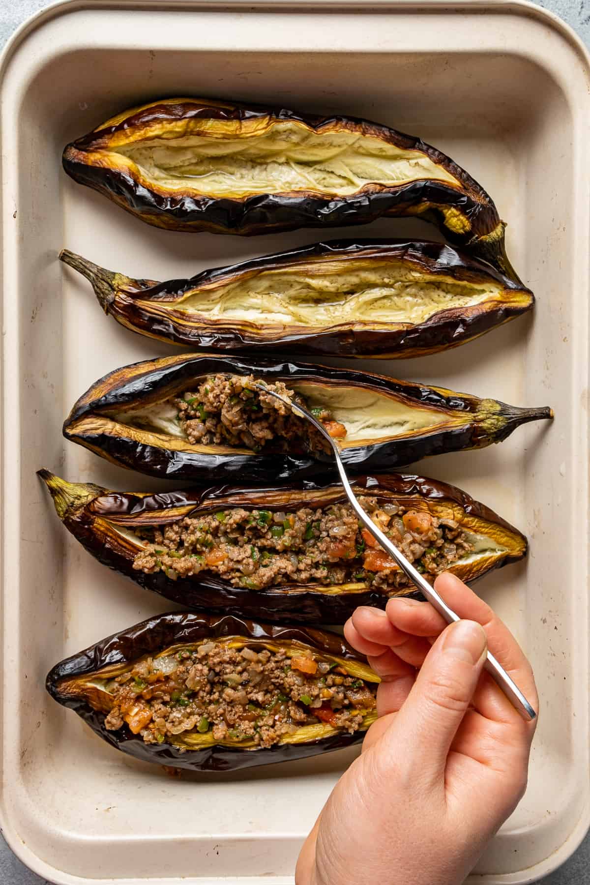 A hand stuffing the roasted whole eggplants with ground beef filling with a spoon.