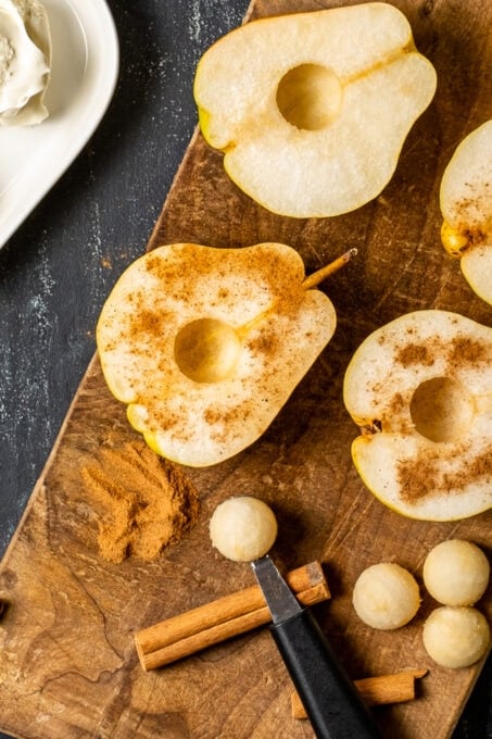 Halved and cored pears with cinnamon powder on them on a wooden board, melon baller and cinnamon sticks on the side.