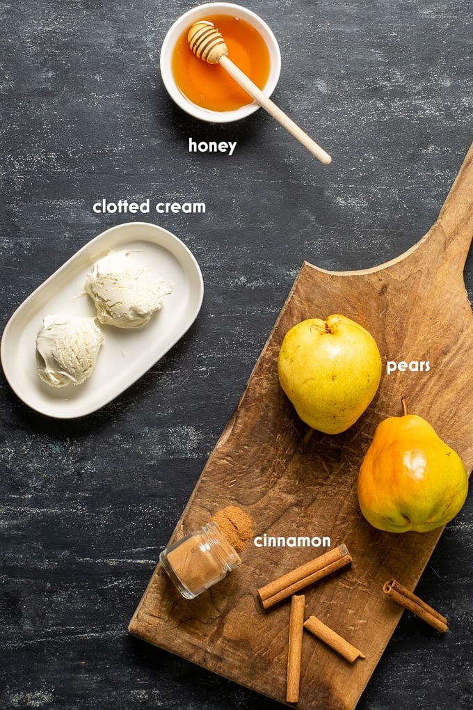 Two pears, cinnamon powder in a mini jar, cinnamon sticks on a wooden board, clotted cream and honey on the side on a black background.