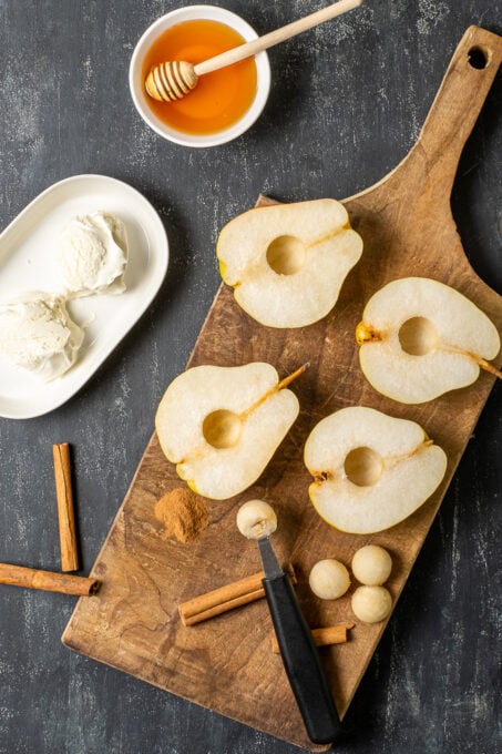 Pears halved and cored on a wooden board, cinnamon sticks, melon baller, clotted cream and honey on the side.